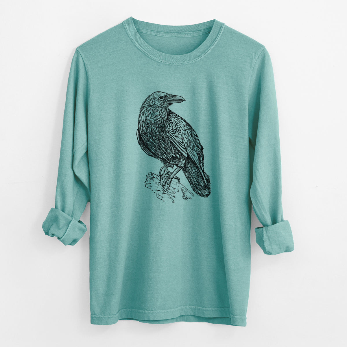 Reef green Comfort Colors long sleeve shirt laid flat with the cuffs rolled, printed with a detailed black ink sketch of a common raven perched on a rock.