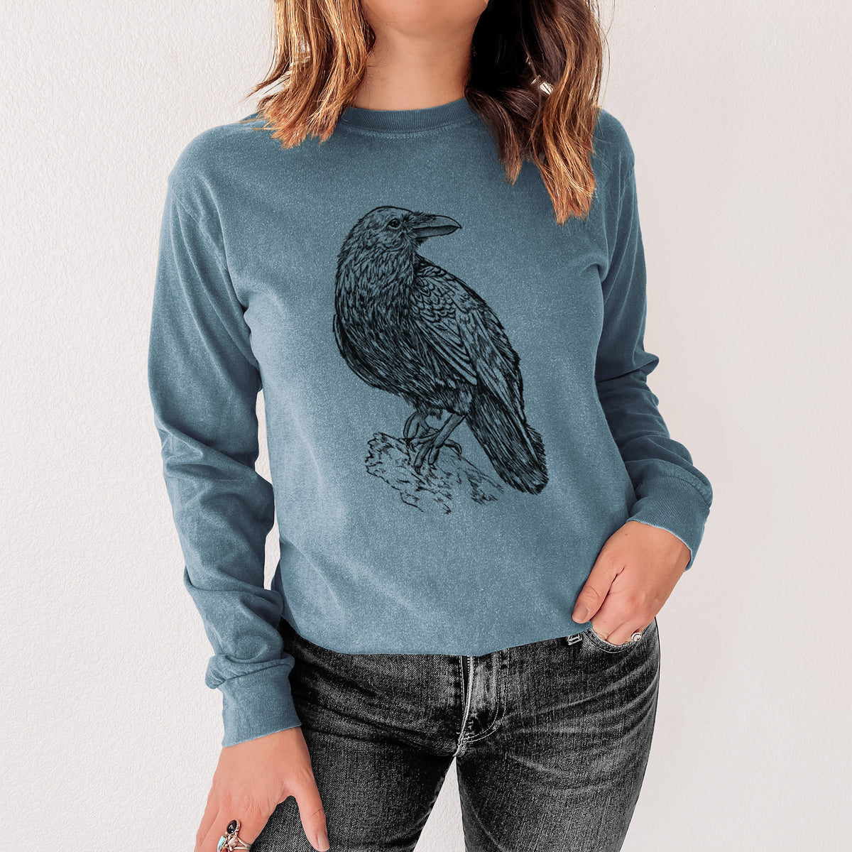 Steel blue Comfort Colors long sleeve shirt printed with a detailed black ink sketch of a common raven perched on a rock, shown in a lifestyle photo.