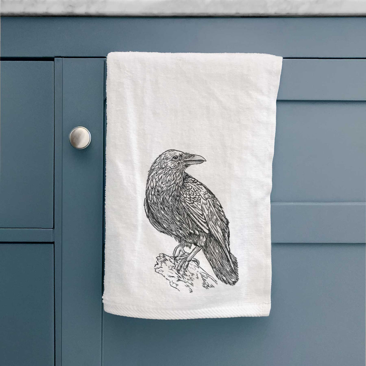 White hand towel printed with a detailed black ink sketch of a common raven perched on a rock, hanging on a cabinet door.