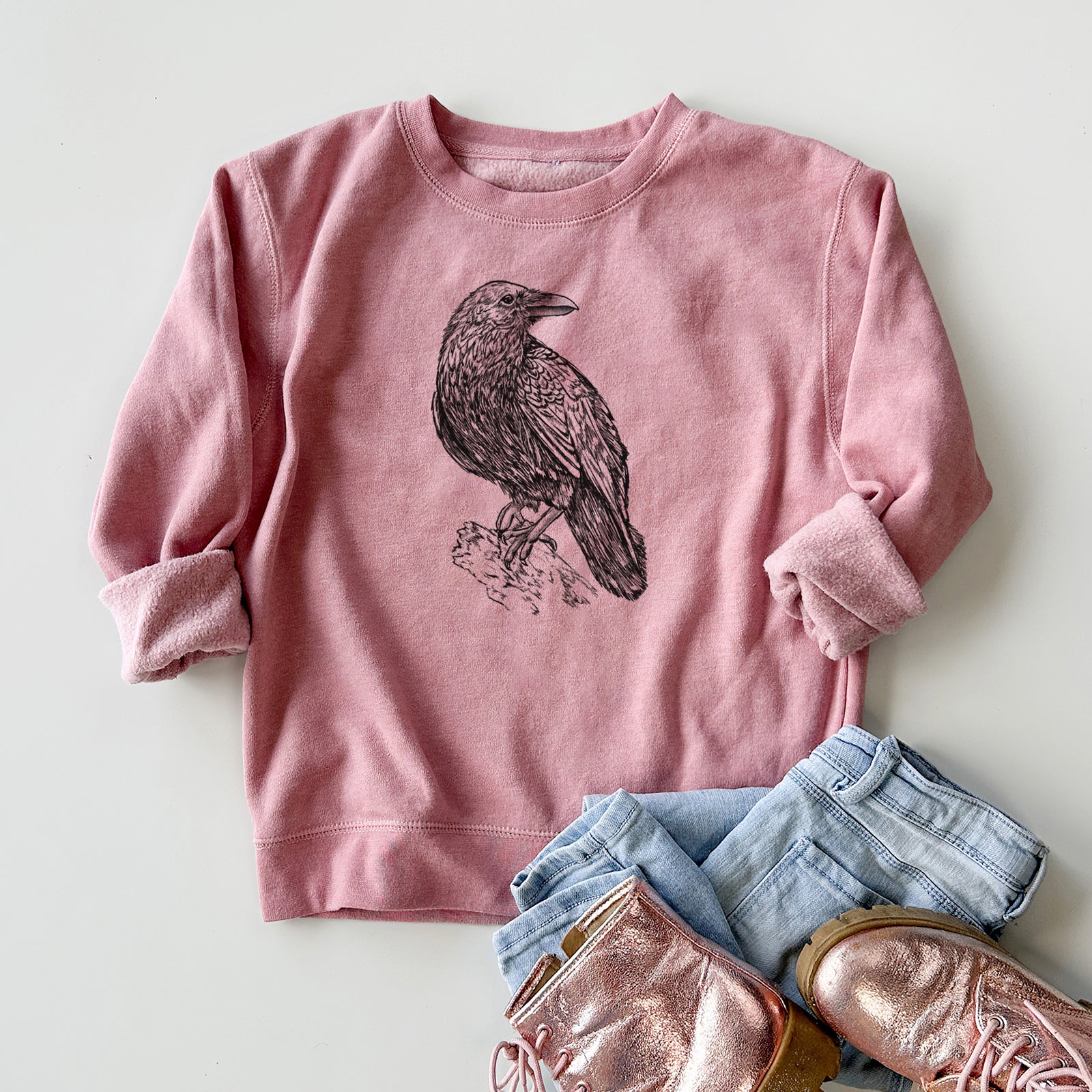 Boy wearing a youth crewneck sweatshirt printed with a detailed black ink sketch of a common raven perched on a rock, outdoors.