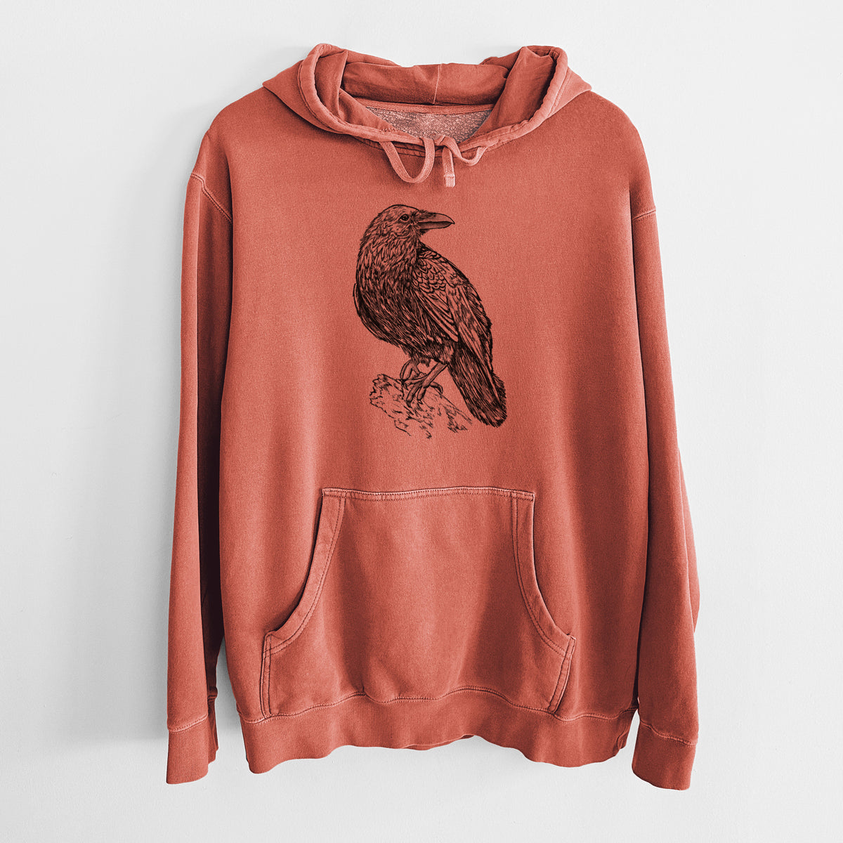 Amber orange pigment dyed hoodie laid flat, printed with a detailed black ink sketch of a common raven perched on a rock.