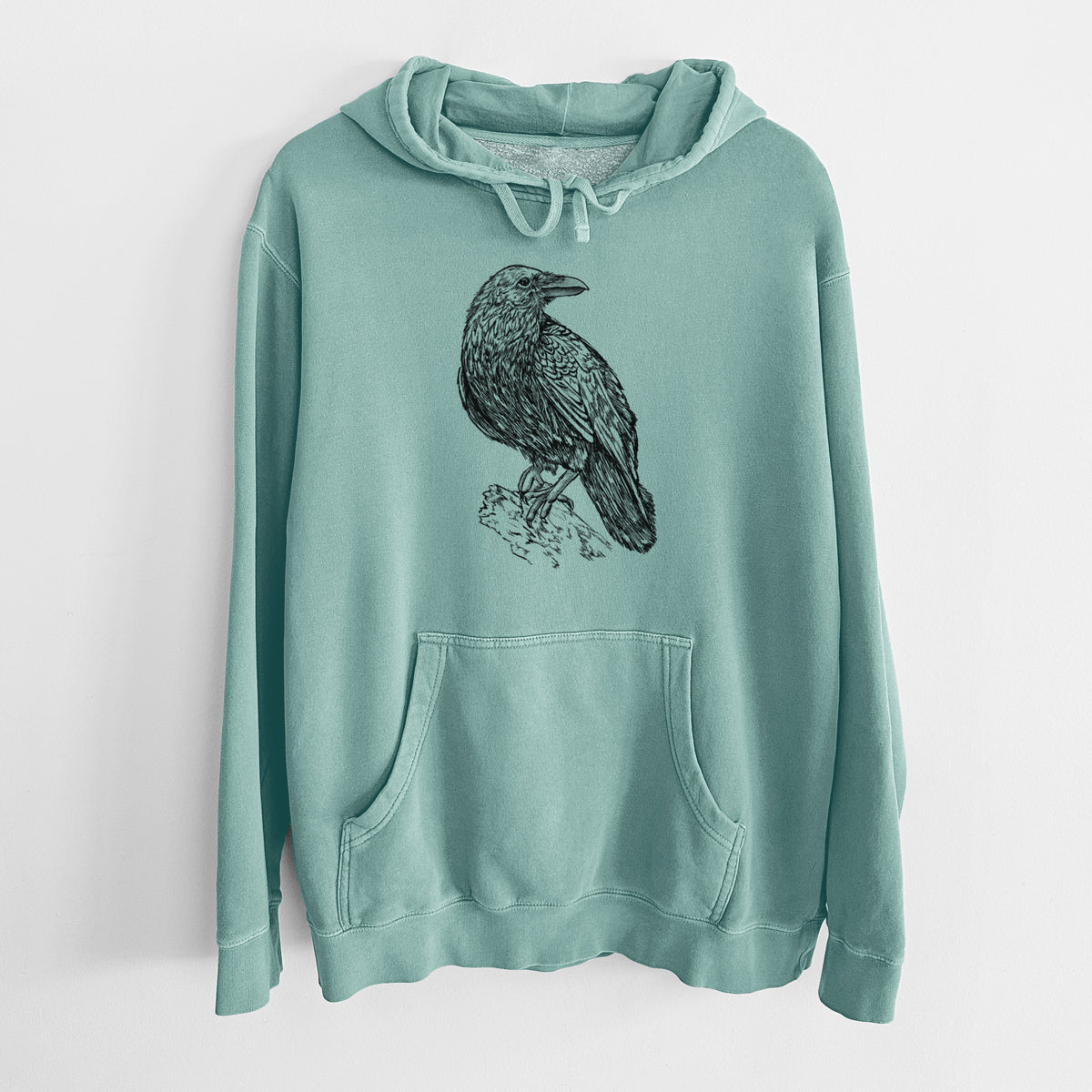 Mint green pigment dyed hoodie laid flat, printed with a detailed black ink sketch of a common raven perched on a rock.