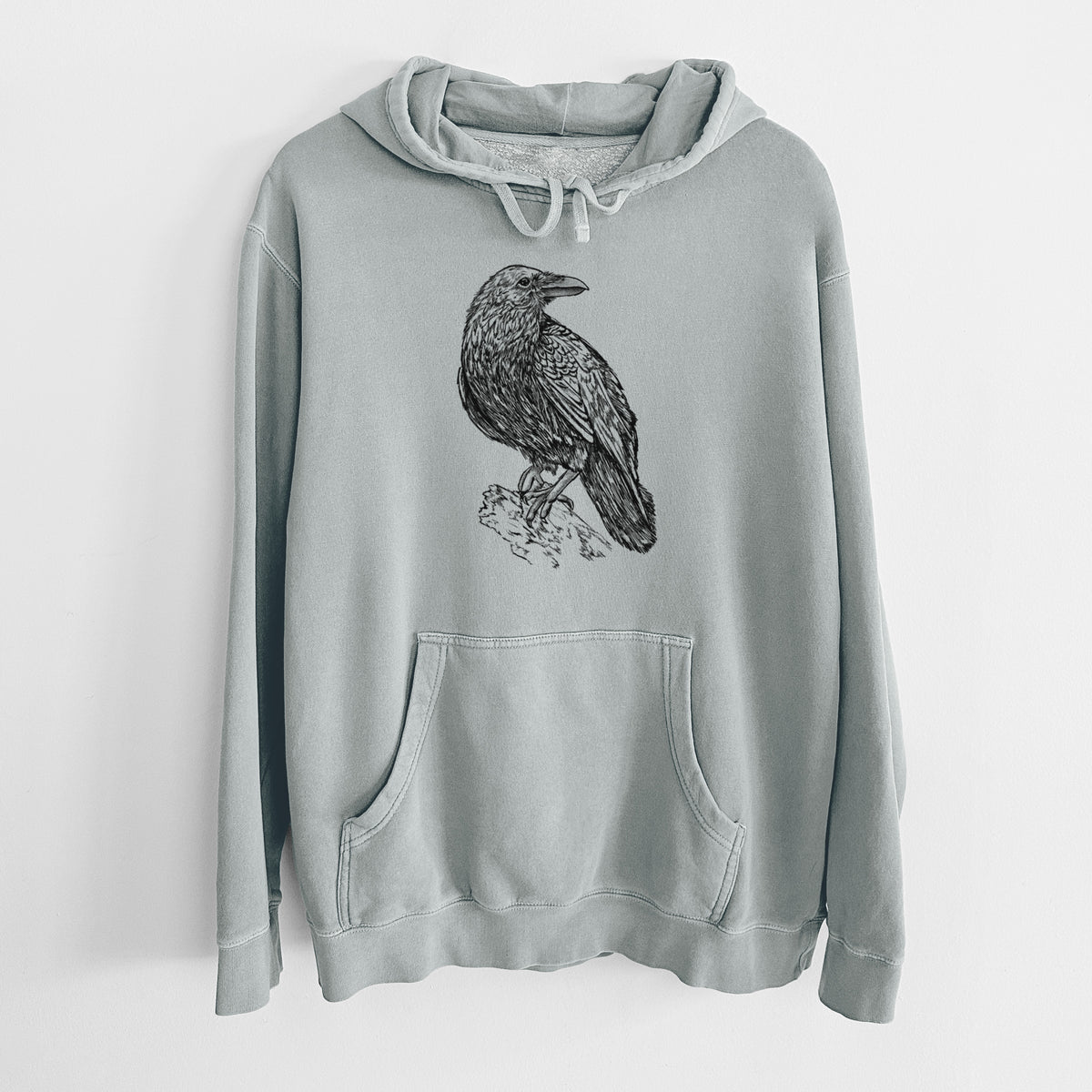 Sage green pigment dyed hoodie laid flat, printed with a detailed black ink sketch of a common raven perched on a rock.