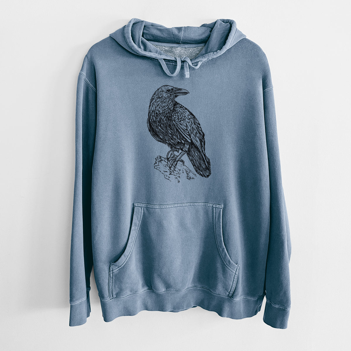 Slate blue pigment dyed hoodie laid flat, printed with a detailed black ink sketch of a common raven perched on a rock.