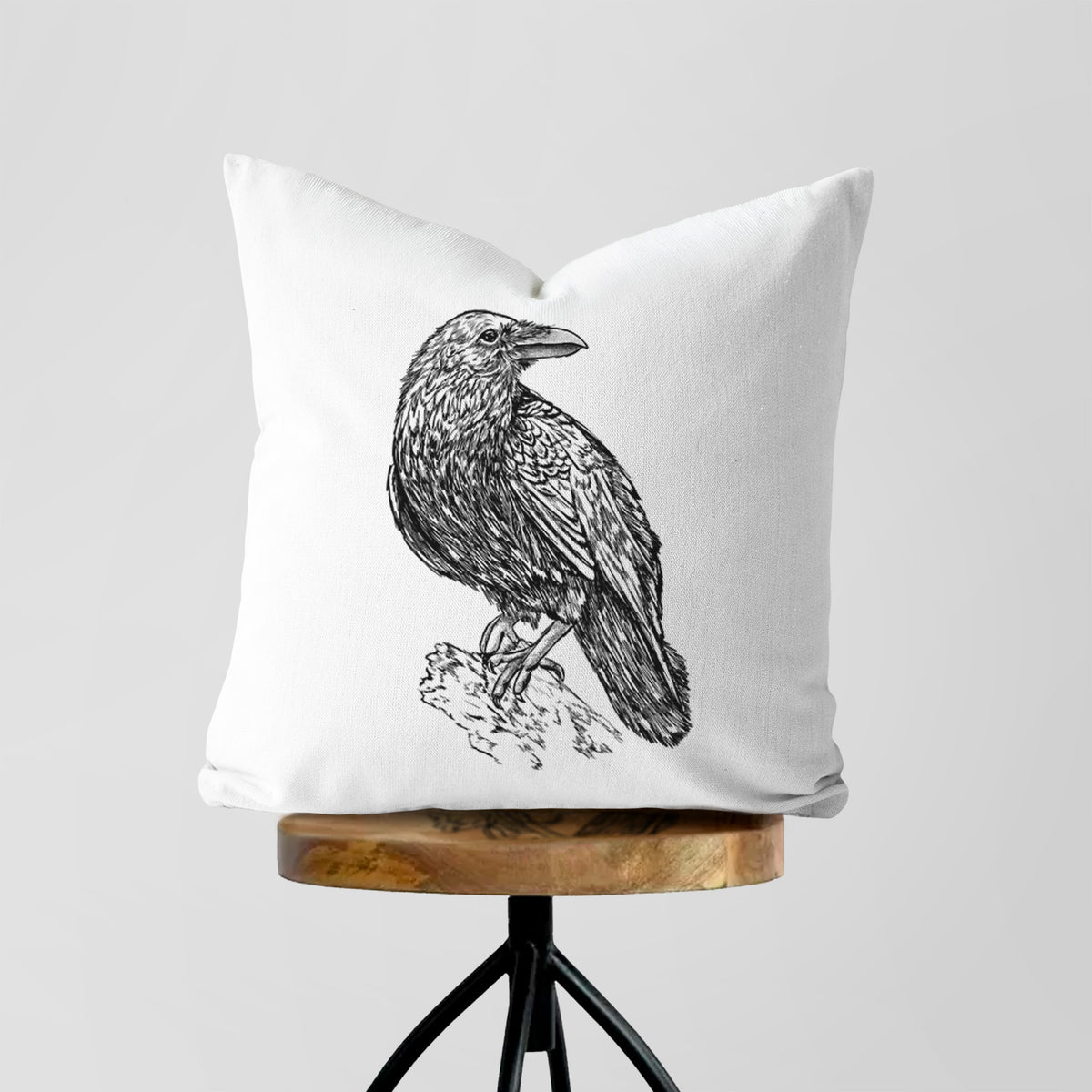 White pillow cover printed with a detailed black ink sketch of a common raven perched on a rock, on a wooden stool.