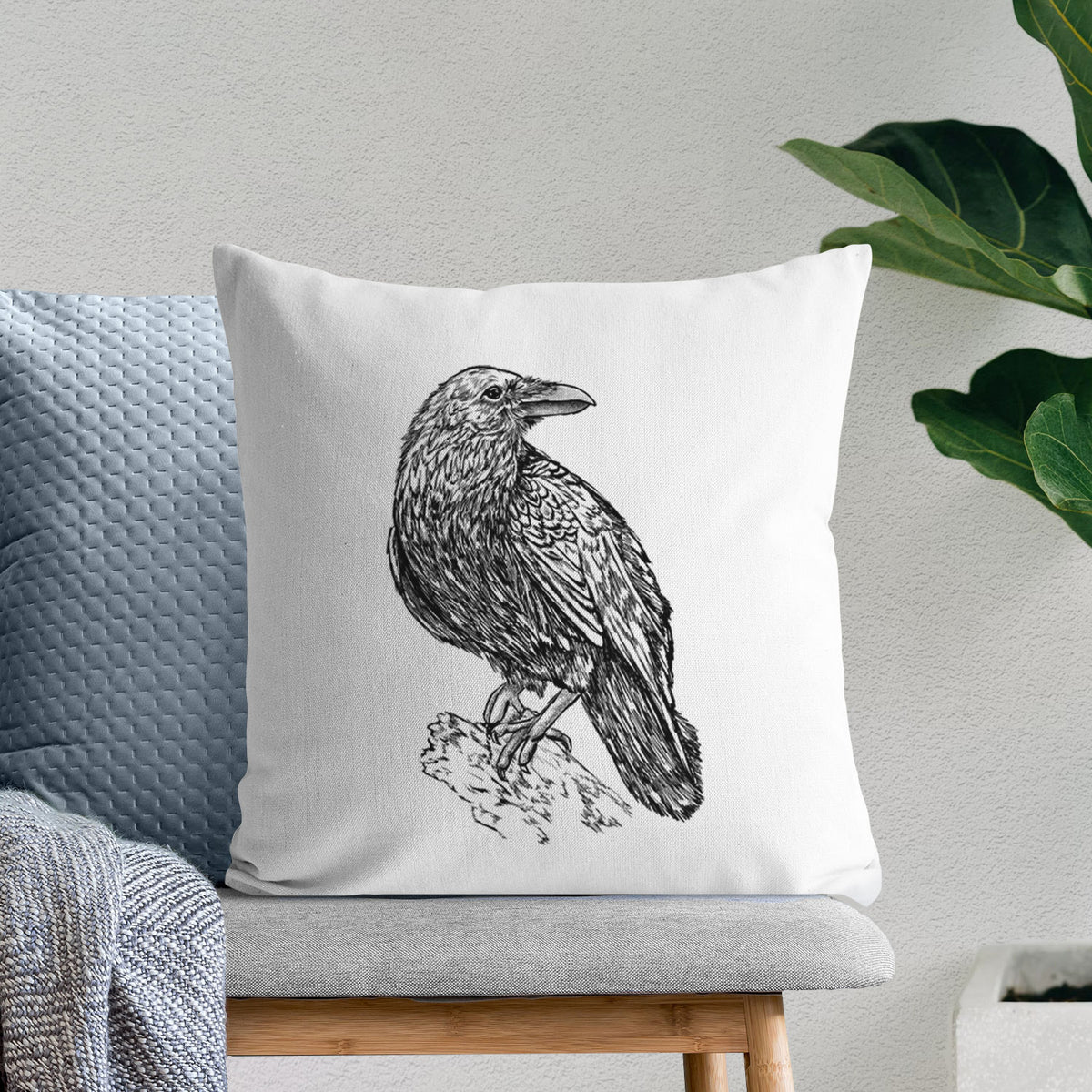 White pillow cover printed with a detailed black ink sketch of a common raven perched on a rock, on a bench beside plants.