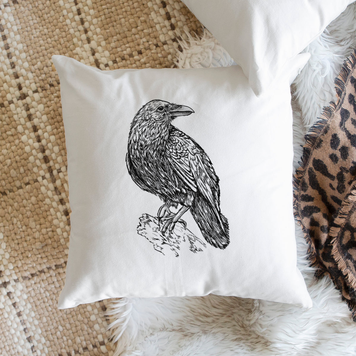 White pillow cover printed with a detailed black ink sketch of a common raven perched on a rock, on a woven chair.