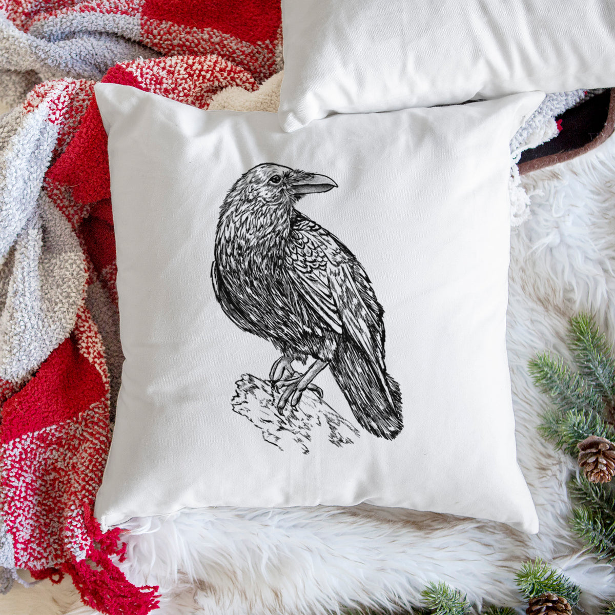 White pillow cover printed with a detailed black ink sketch of a common raven perched on a rock, on a woven chair.