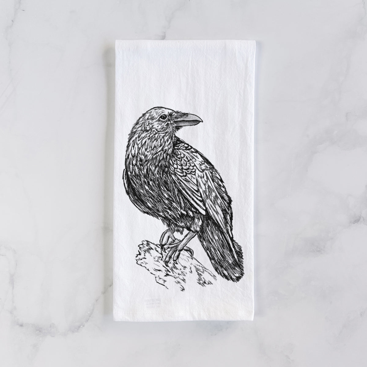 White cotton tea towel laid flat on marble, printed with a detailed black ink sketch of a common raven perched on a rock.