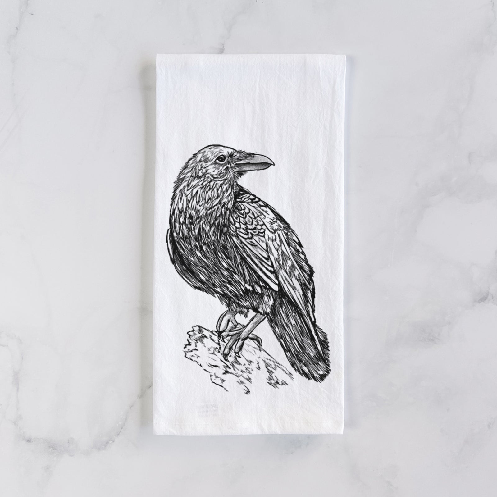 White cotton tea towel printed with a detailed black ink sketch of a common raven perched on a rock, on a marble counter beside a round wooden cutting board and greenery.