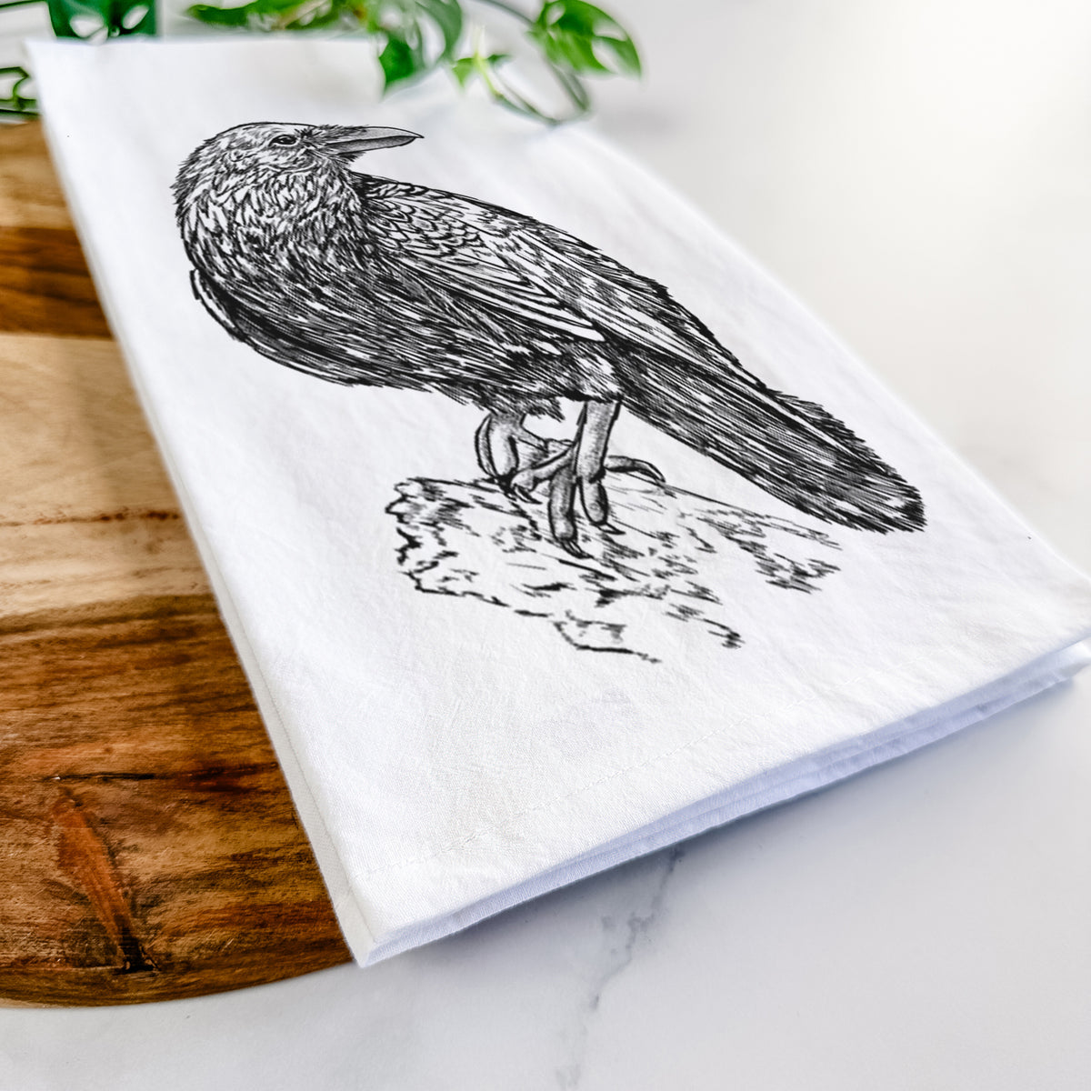 Close-up of a folded white tea towel on a wooden board, showing a detailed black ink sketch of a common raven perched on a rock.