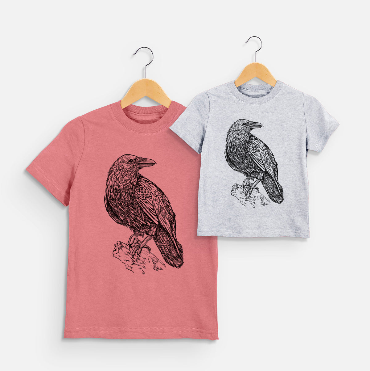 Two youth t-shirts in different colors on hangers, printed with a detailed black ink sketch of a common raven perched on a rock.