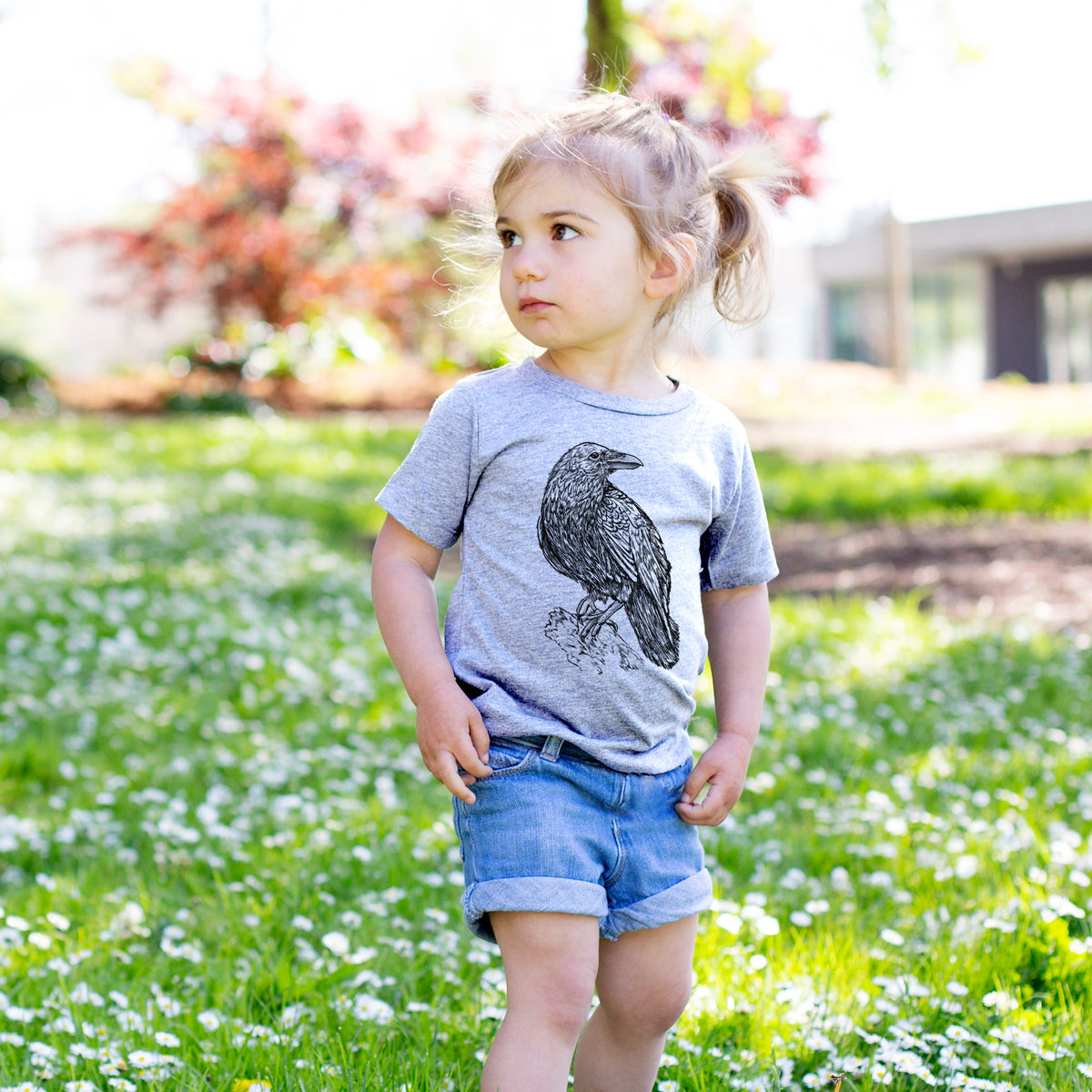 Toddler girl walking outdoors wearing a t-shirt printed with a detailed black ink sketch of a common raven perched on a rock.
