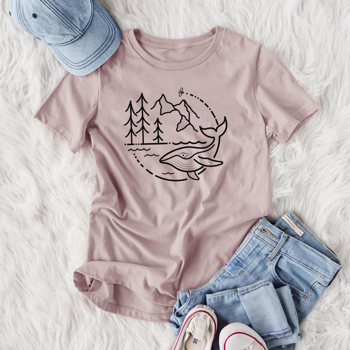 Women's relaxed fit cotton t-shirt printed with a black line art circle connecting a mountain range, pine trees, a bee and a whale swimming in ocean waves, styled flat on a white faux fur rug with jeans.