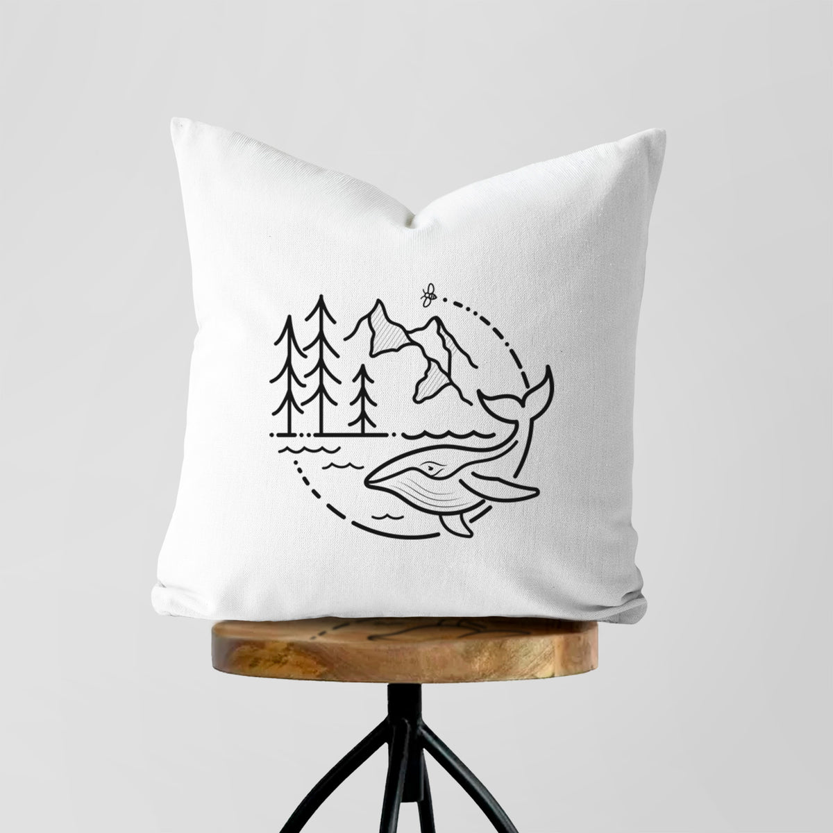 White pillow cover printed with a black line art circle connecting a mountain range, pine trees, a bee and a whale swimming in ocean waves, on a wooden stool.