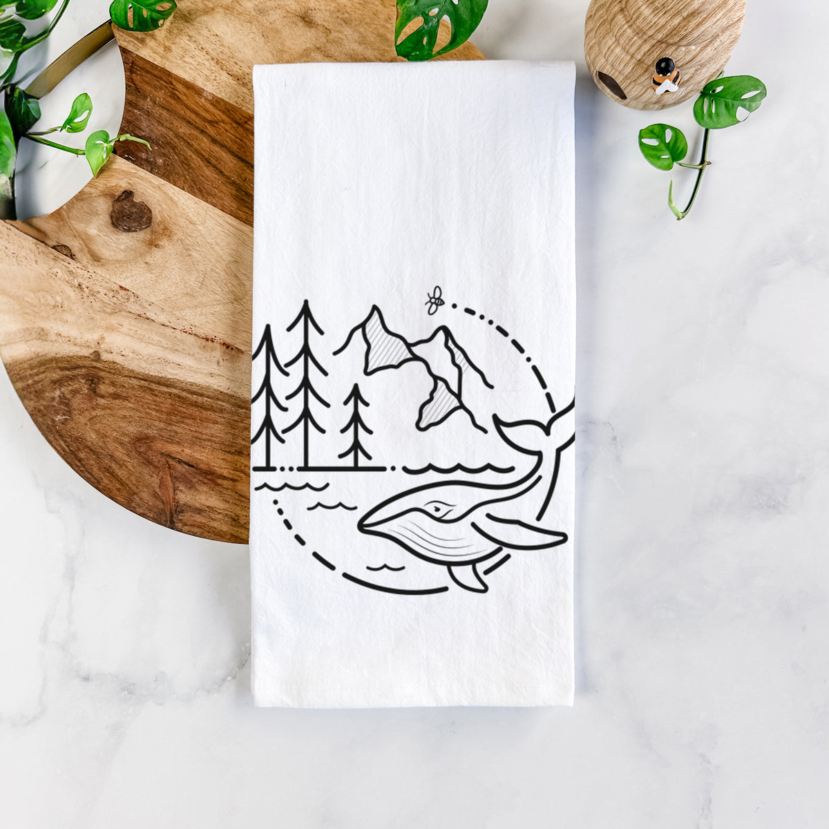 White cotton tea towel printed with a black line art circle connecting a mountain range, pine trees, a bee and a whale swimming in ocean waves, on a marble counter beside a round wooden cutting board and greenery.