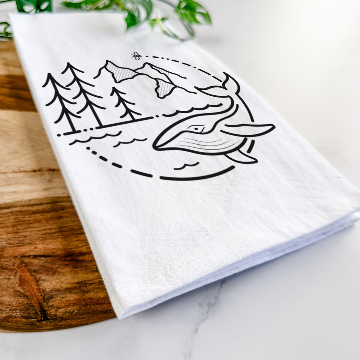 Close-up of a folded white tea towel on a wooden board, showing a black line art circle connecting a mountain range, pine trees, a bee and a whale swimming in ocean waves.