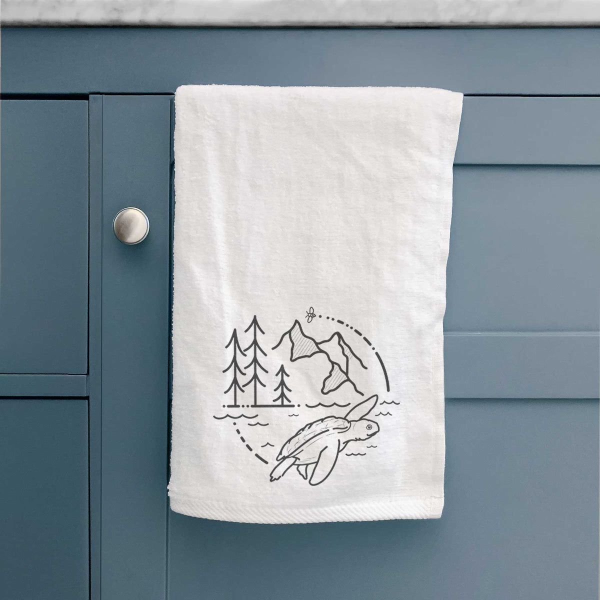 White hand towel printed with a black line art circle connecting pine trees, mountains, a bee and a Kemp's ridley sea turtle swimming, hanging on a cabinet door.