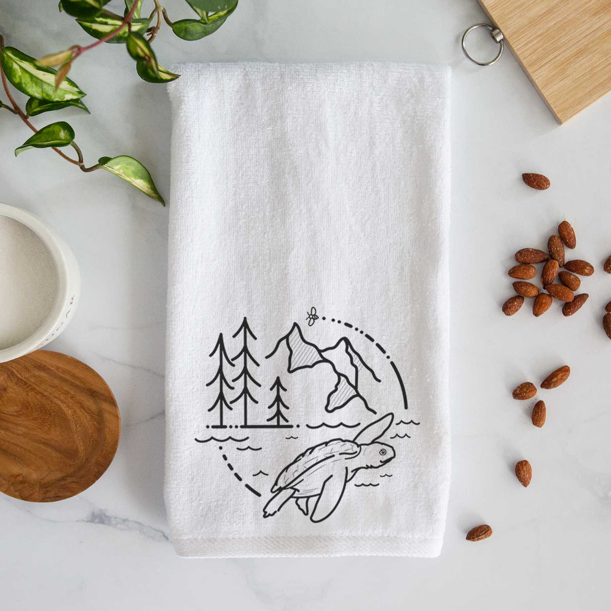 White hand towel printed with a black line art circle connecting pine trees, mountains, a bee and a Kemp's ridley sea turtle swimming.