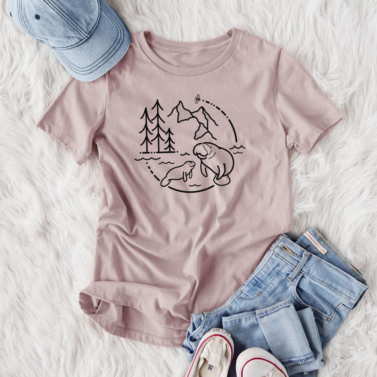 Women's relaxed fit cotton t-shirt printed with a black line art circle connecting pine trees, mountains, a bee and two manatees swimming in water, styled flat on a white faux fur rug with jeans.