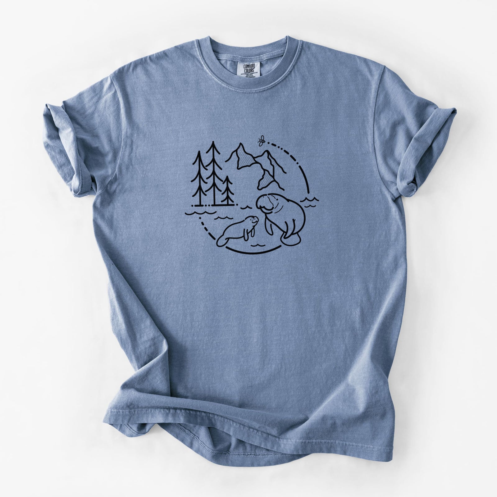 Blue jean Comfort Colors t-shirt laid flat, printed with a black line art circle connecting pine trees, mountains, a bee and two manatees swimming in water.