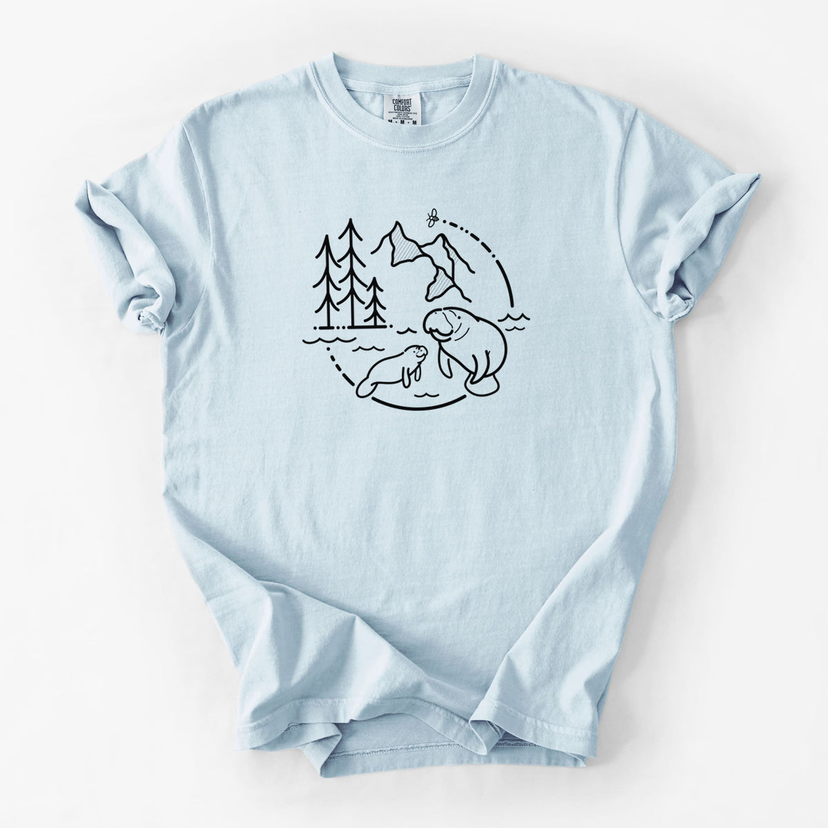 Chambray blue Comfort Colors t-shirt laid flat, printed with a black line art circle connecting pine trees, mountains, a bee and two manatees swimming in water.