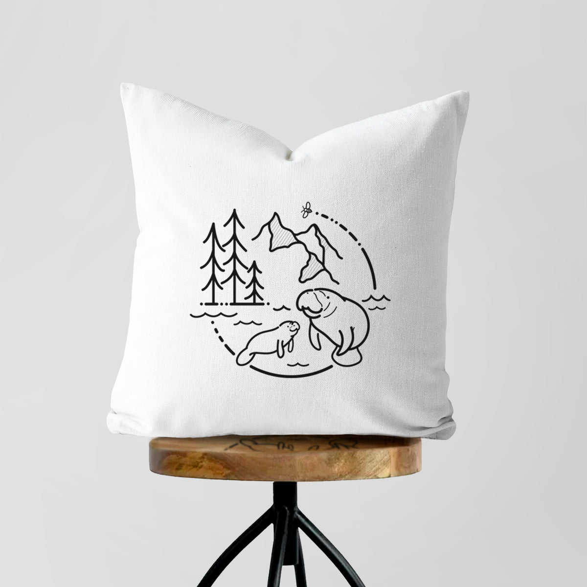 White pillow cover printed with a black line art circle connecting pine trees, mountains, a bee and two manatees swimming in water, on a wooden stool.