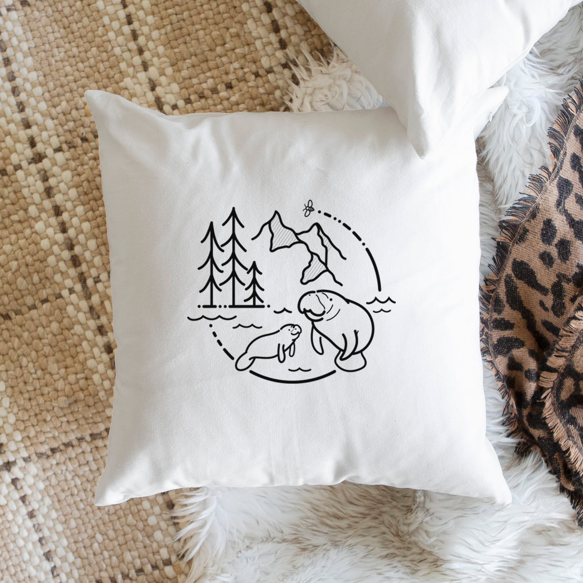 White pillow cover printed with a black line art circle connecting pine trees, mountains, a bee and two manatees swimming in water, on a woven chair.