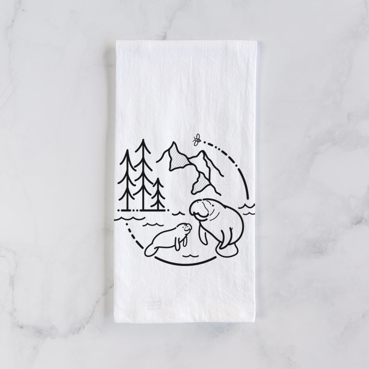 White cotton tea towel laid flat on marble, printed with a black line art circle connecting pine trees, mountains, a bee and two manatees swimming in water.