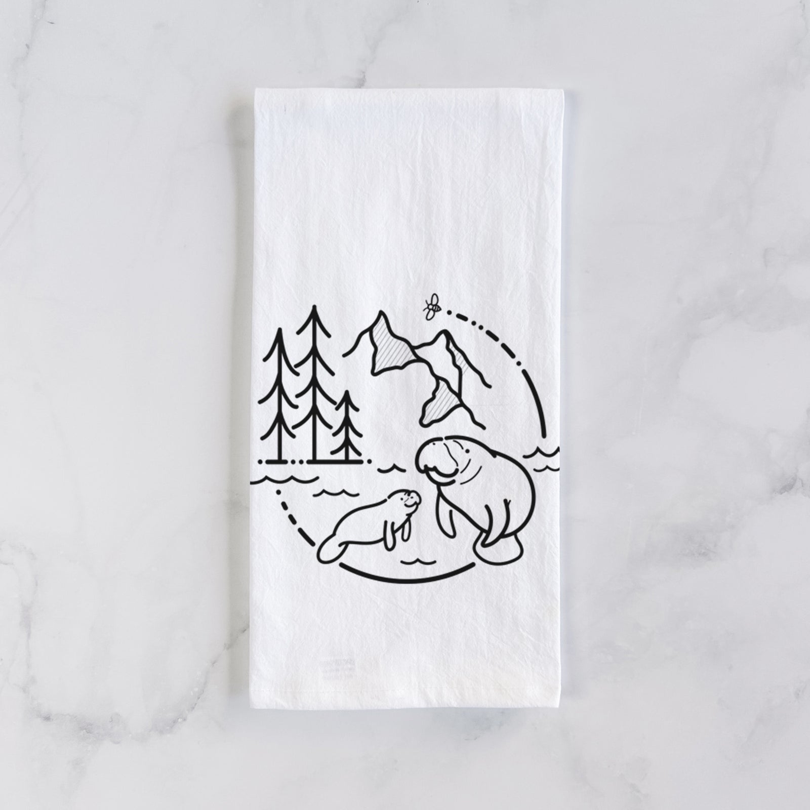 White cotton tea towel printed with a black line art circle connecting pine trees, mountains, a bee and two manatees swimming in water, on a marble counter beside a round wooden cutting board and greenery.