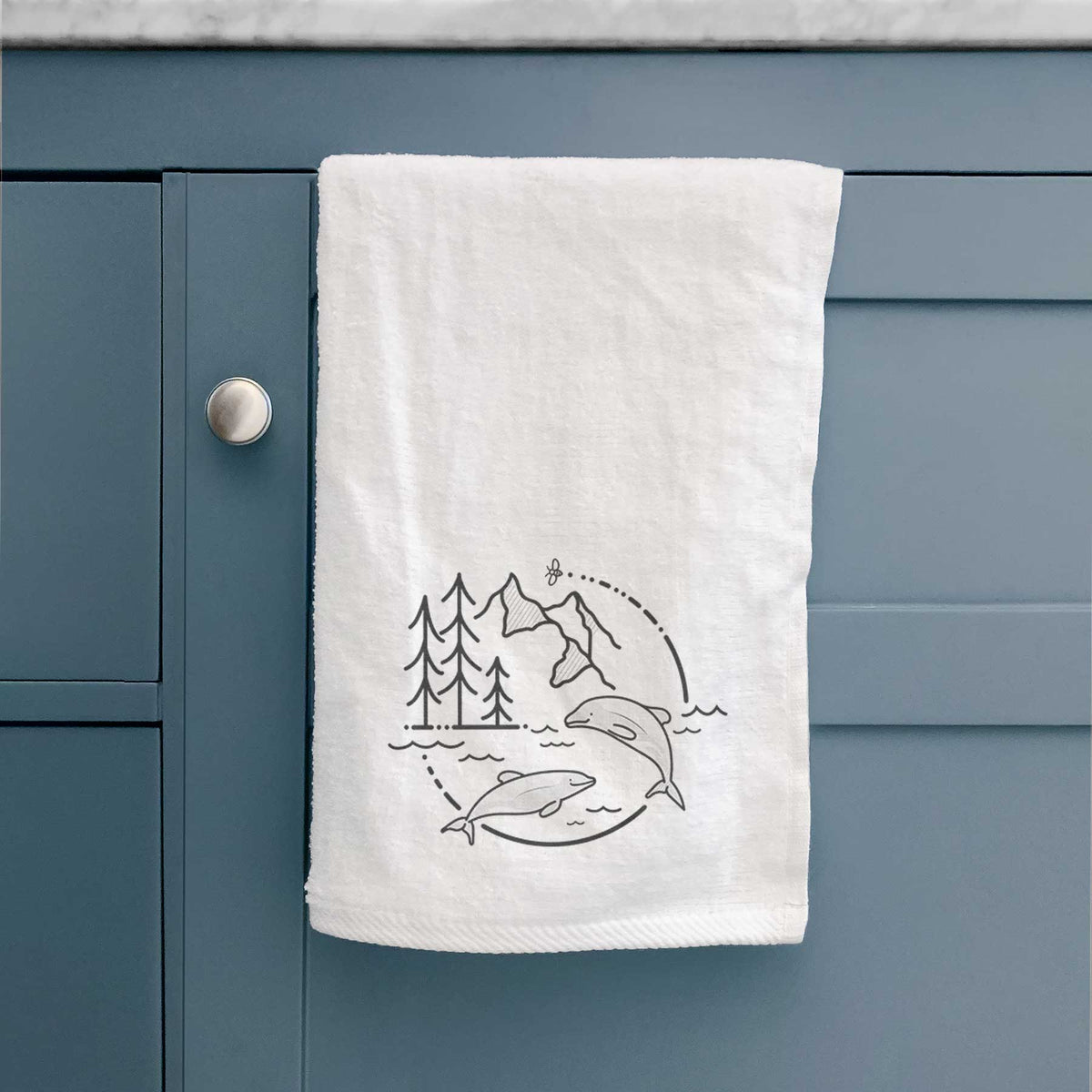 White hand towel printed with a black line art circle connecting pine trees, mountains, a bee and two Maui dolphins leaping from the waves, hanging on a cabinet door.