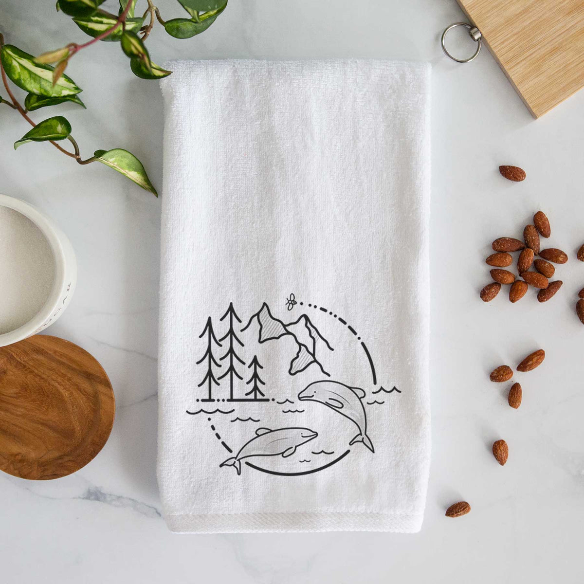 White hand towel printed with a black line art circle connecting pine trees, mountains, a bee and two Maui dolphins leaping from the waves.