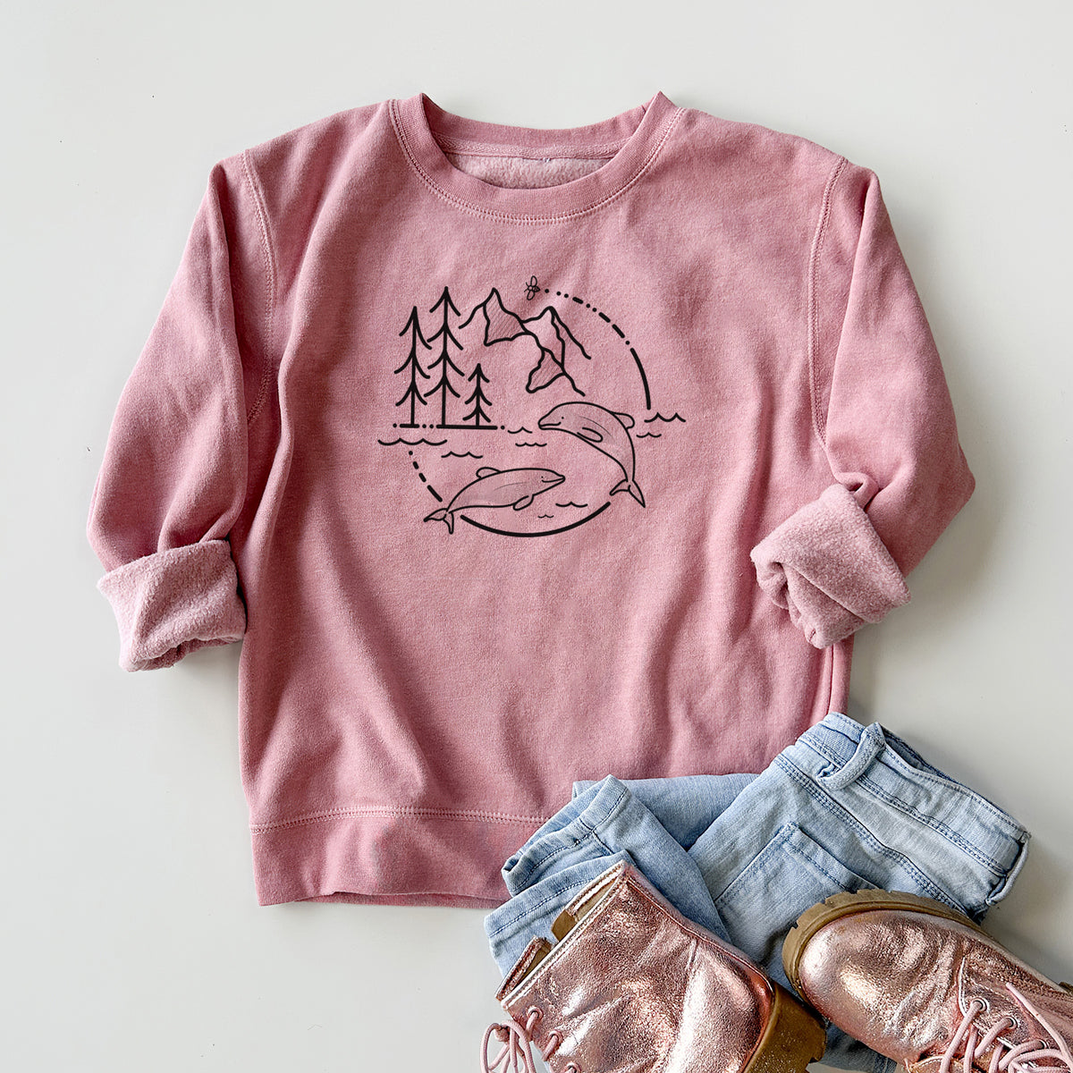 Youth crewneck sweatshirt printed with a black line art circle connecting pine trees, mountains, a bee and two Maui dolphins leaping from the waves, styled flat with jeans and boots.