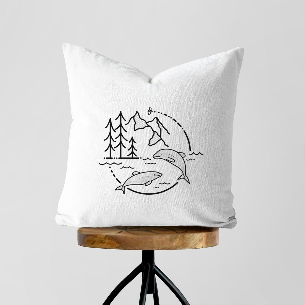 White pillow cover printed with a black line art circle connecting pine trees, mountains, a bee and two Maui dolphins leaping from the waves, on a wooden stool.