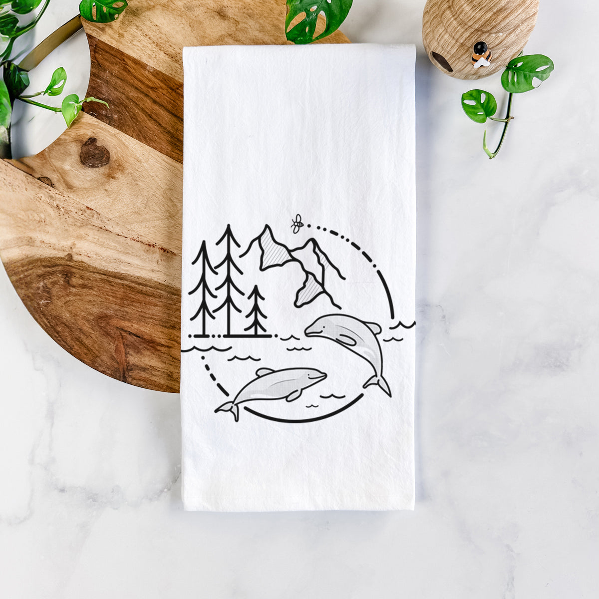 White cotton tea towel printed with a black line art circle connecting pine trees, mountains, a bee and two Maui dolphins leaping from the waves, on a marble counter beside a round wooden cutting board and greenery.