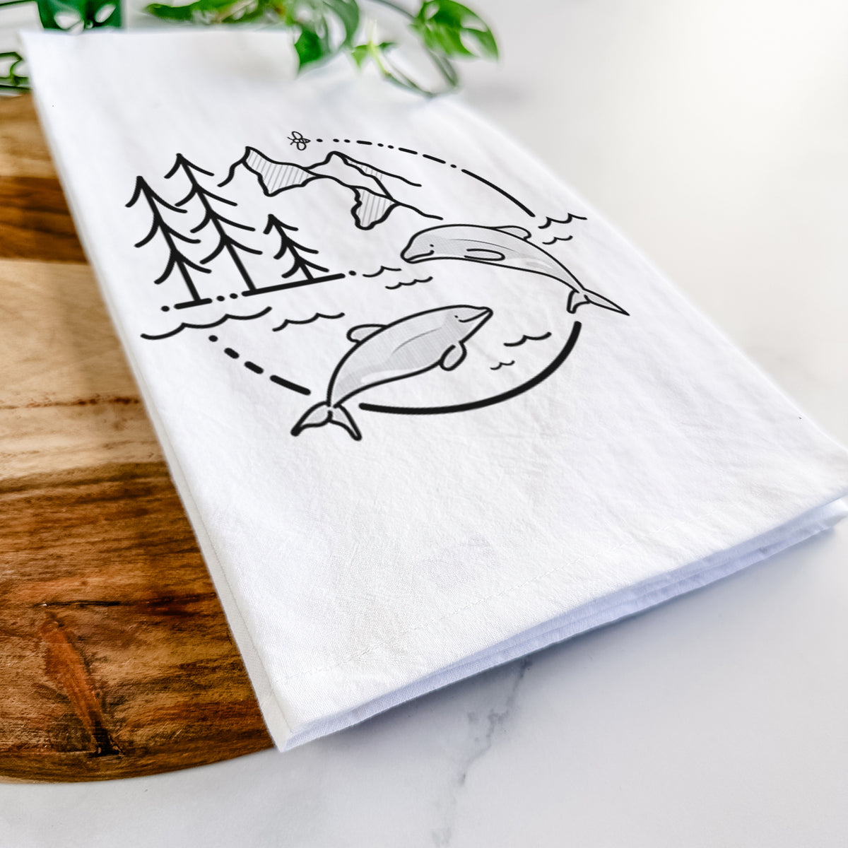 Close-up of a folded white tea towel on a wooden board, showing a black line art circle connecting pine trees, mountains, a bee and two Maui dolphins leaping from the waves.