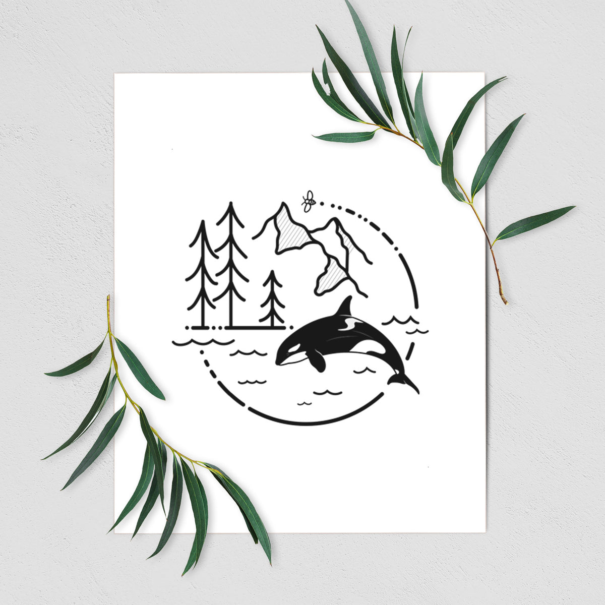 Art print of a black line art circle connecting pine trees, mountains, a bee and an orca leaping from ocean waves, shown with eucalyptus leaves.
