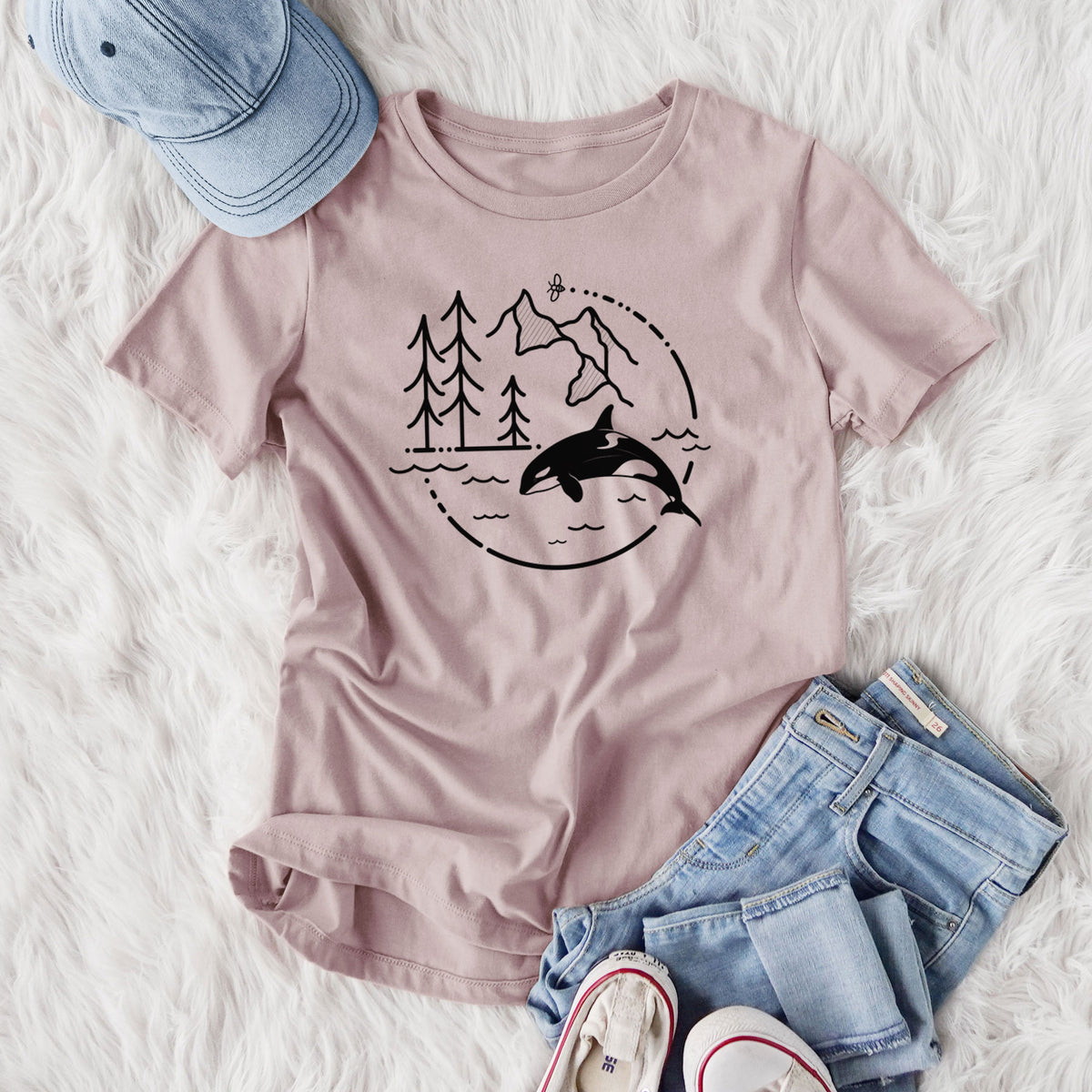 Women's relaxed fit cotton t-shirt printed with a black line art circle connecting pine trees, mountains, a bee and an orca leaping from ocean waves, styled flat on a white faux fur rug with jeans.