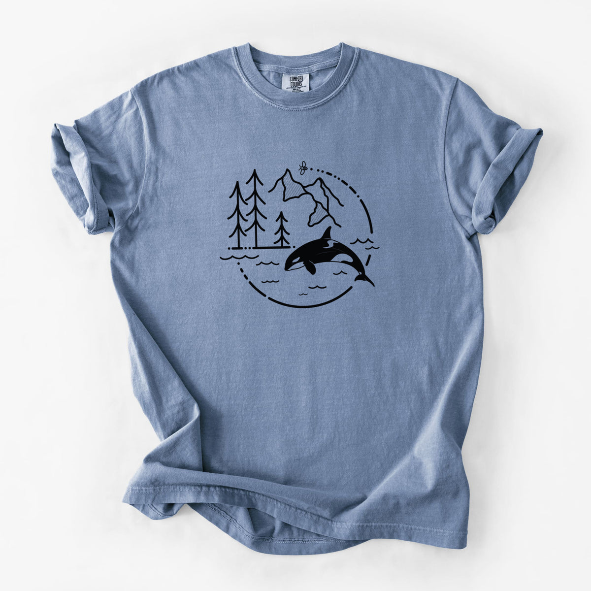 Blue jean Comfort Colors t-shirt laid flat, printed with a black line art circle connecting pine trees, mountains, a bee and an orca leaping from ocean waves.