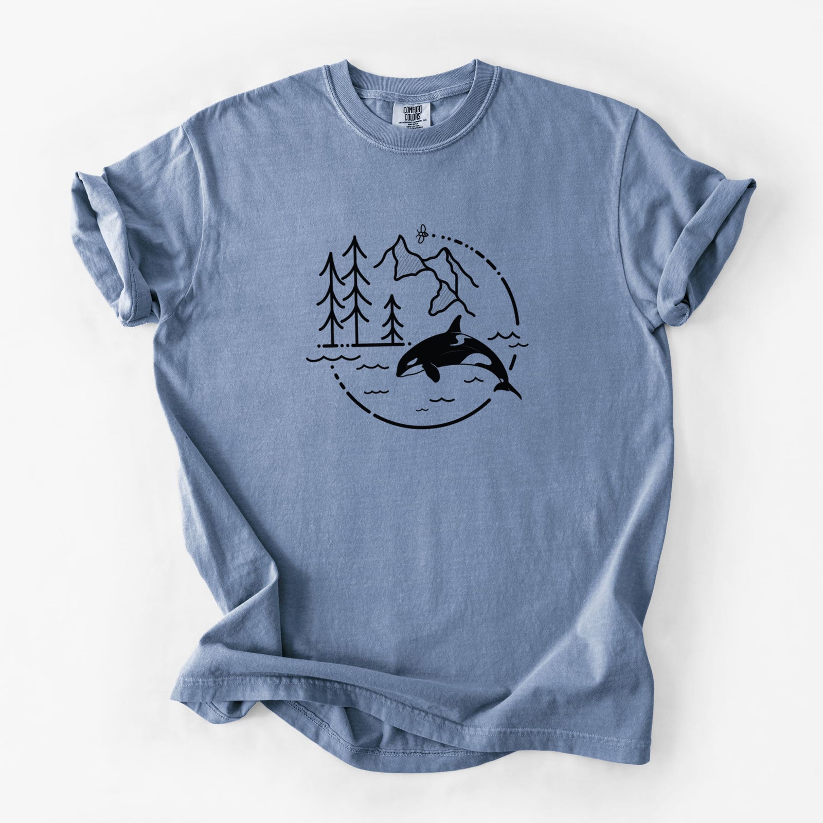 Blue jean Comfort Colors t-shirt laid flat, printed with a black line art circle connecting pine trees, mountains, a bee and an orca leaping from ocean waves.