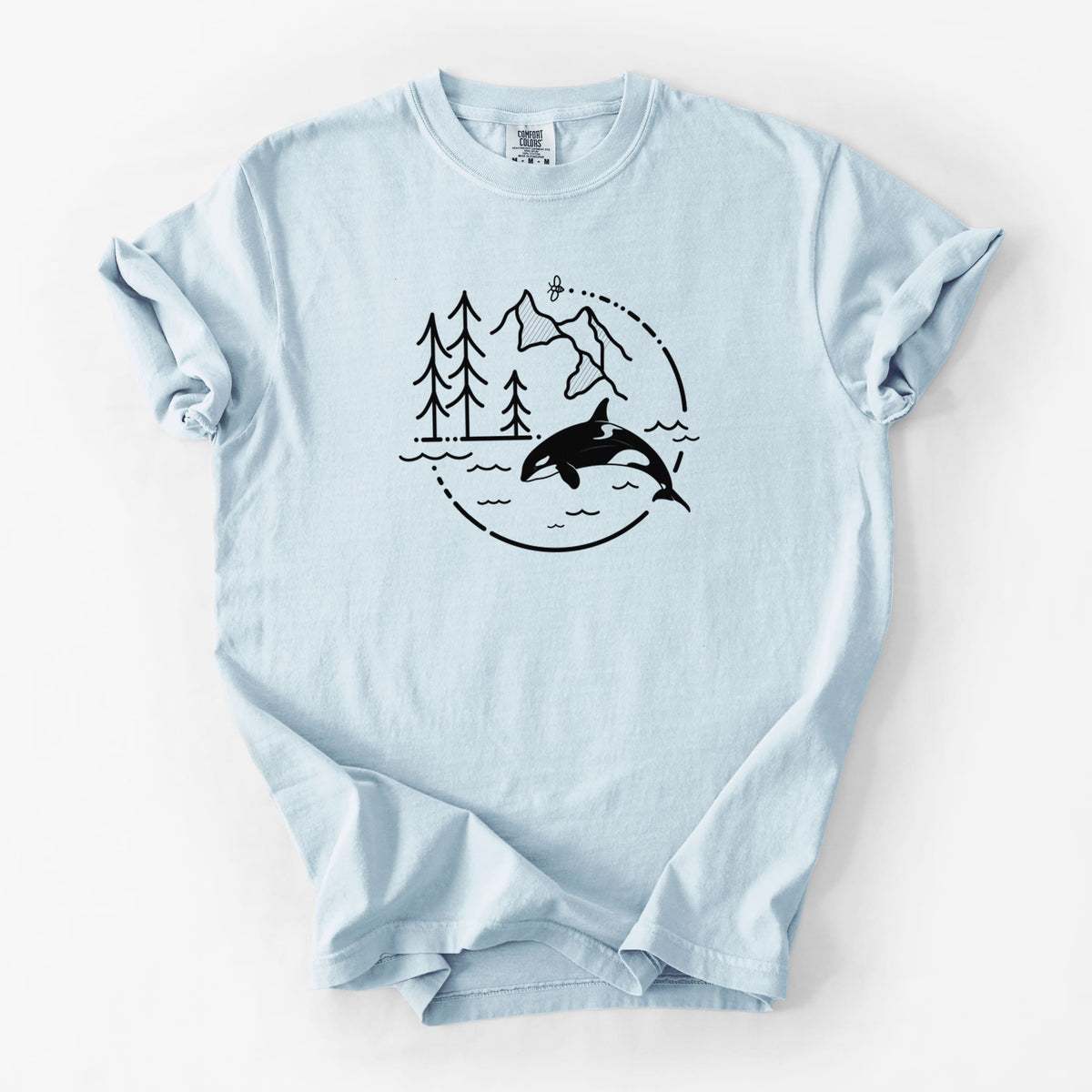 Chambray blue Comfort Colors t-shirt laid flat, printed with a black line art circle connecting pine trees, mountains, a bee and an orca leaping from ocean waves.