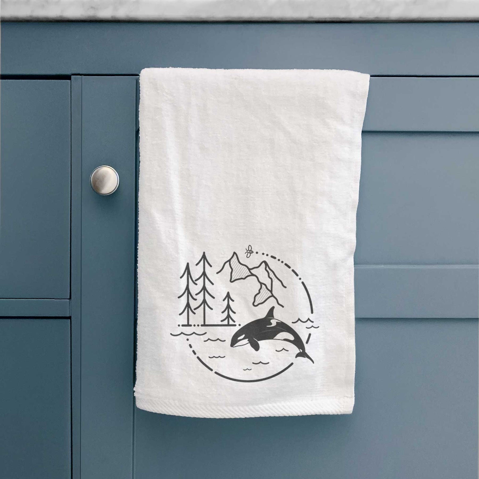 White hand towel printed with a black line art circle connecting pine trees, mountains, a bee and an orca leaping from ocean waves.