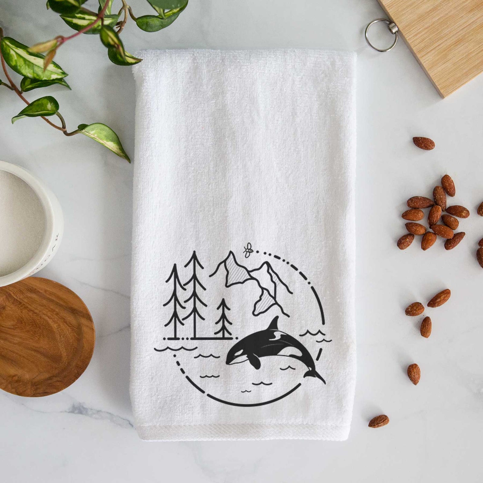 White hand towel printed with a black line art circle connecting pine trees, mountains, a bee and an orca leaping from ocean waves.