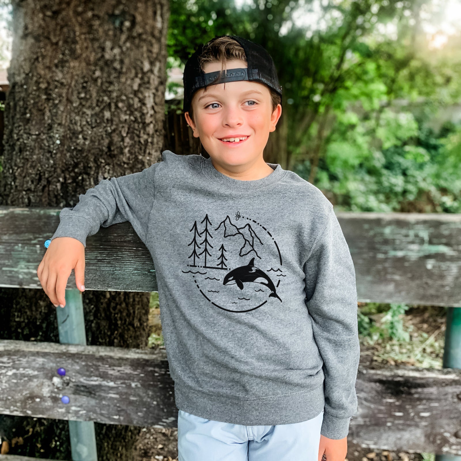 Boy wearing a youth crewneck sweatshirt printed with a black line art circle connecting pine trees, mountains, a bee and an orca leaping from ocean waves, outdoors.