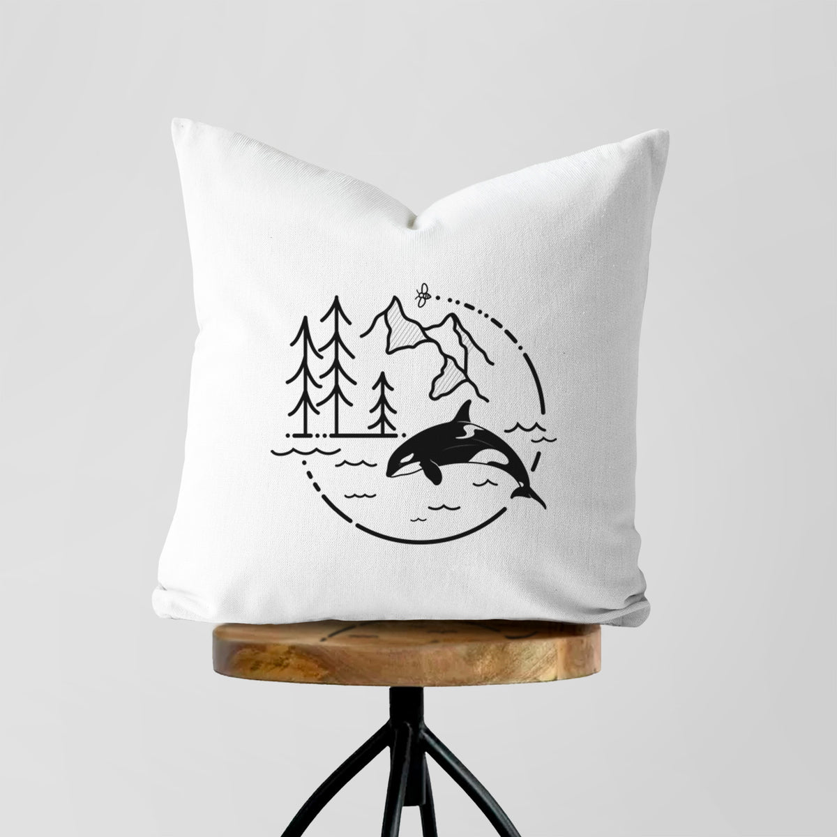 White pillow cover printed with a black line art circle connecting pine trees, mountains, a bee and an orca leaping from ocean waves, on a wooden stool.