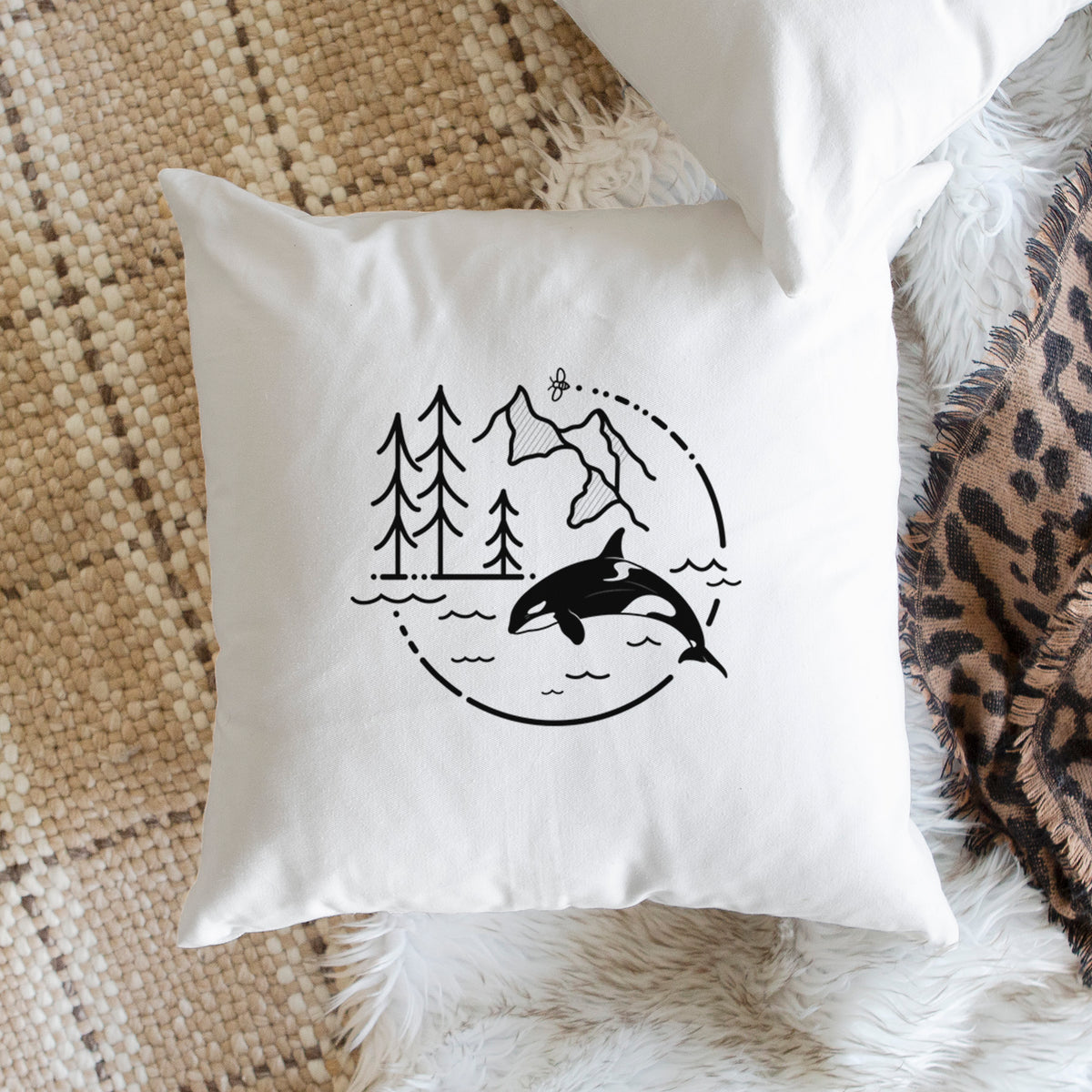 White pillow cover printed with a black line art circle connecting pine trees, mountains, a bee and an orca leaping from ocean waves, on a woven chair.