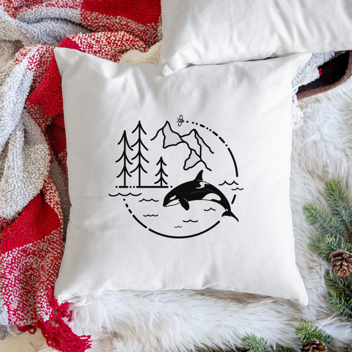 White pillow cover printed with a black line art circle connecting pine trees, mountains, a bee and an orca leaping from ocean waves, on a woven chair.