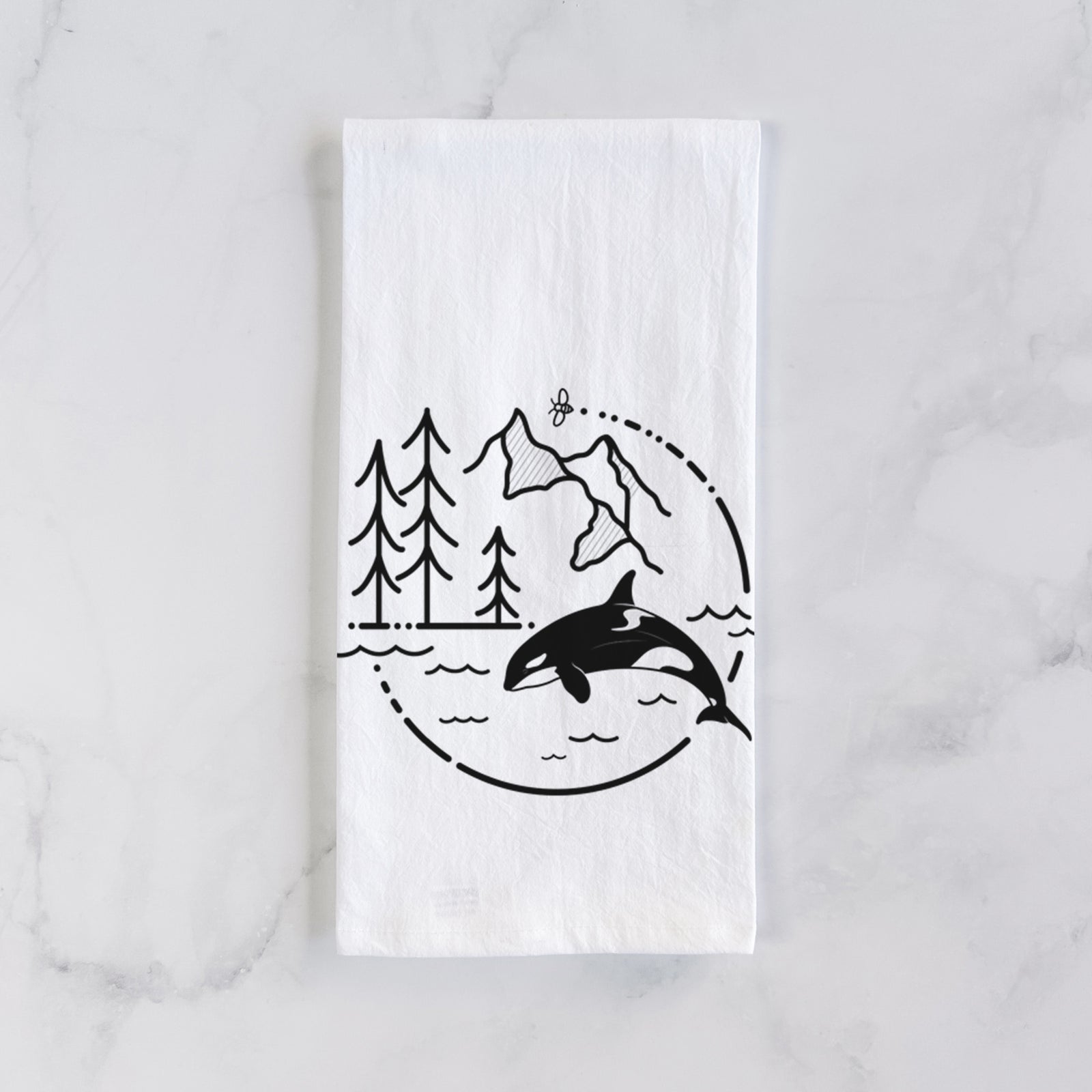 White cotton tea towel printed with a black line art circle connecting pine trees, mountains, a bee and an orca leaping from ocean waves, on a marble counter beside a round wooden cutting board and greenery.