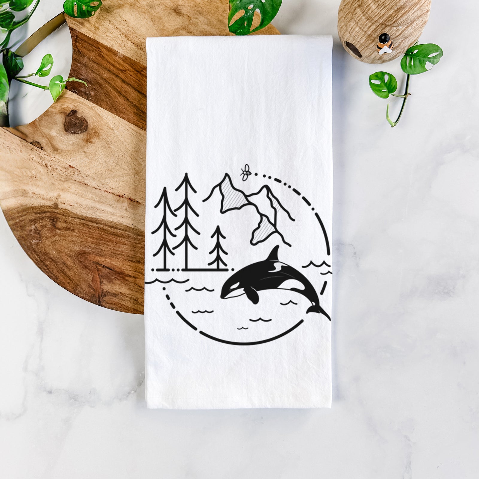 White cotton tea towel printed with a black line art circle connecting pine trees, mountains, a bee and an orca leaping from ocean waves, on a marble counter beside a round wooden cutting board and greenery.