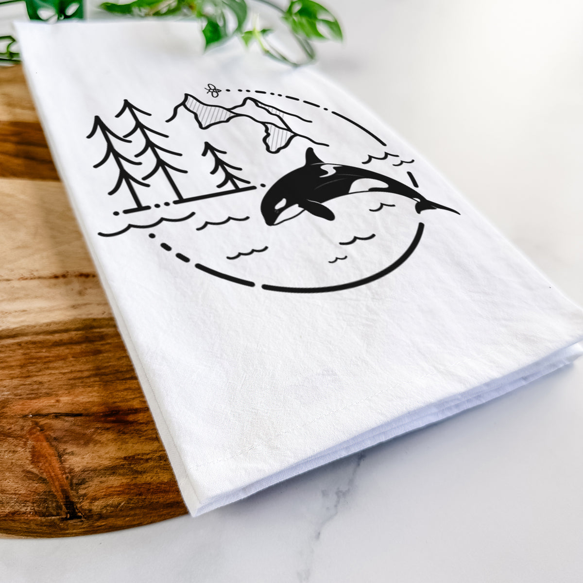 Close-up of a folded white tea towel on a wooden board, showing a black line art circle connecting pine trees, mountains, a bee and an orca leaping from ocean waves.